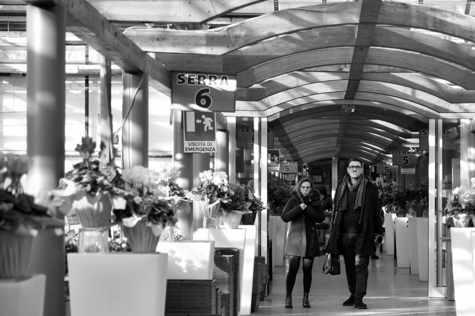 A black and white photograph of an indoor shopping centre corridor with curved, translucent ceiling panels allowing natural light to illuminate the space. Along the corridor, there are large white pla