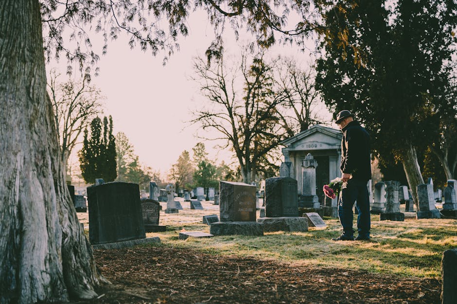 A man dressed in a dark suit and black hat is standing in a cemetery, holding a bouquet of pink and red flowers. Surrounding him are numerous gravestones and memorials made of stone, varying in size a