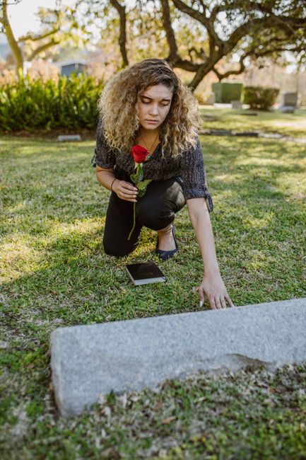 A young woman with curly blonde hair kneeling on a grassy area, placing a red rose on a gravestone during daylight hours. She is dressed in a dark, patterned top and black pants, holding a single rose