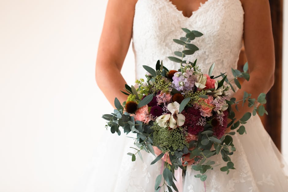 A bride dressed in a lace wedding gown holds a large, vibrant floral bouquet featuring pink, purple, white, and deep burgundy blooms. The arrangement includes roses, hydrangeas, and freesias, accented