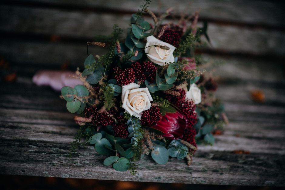 A floral arrangement featuring cream rose blooms with soft, tightly layered petals, complemented by deep red flowers and sprigs of small, dark red berries. The bouquet includes a variety of greenery s