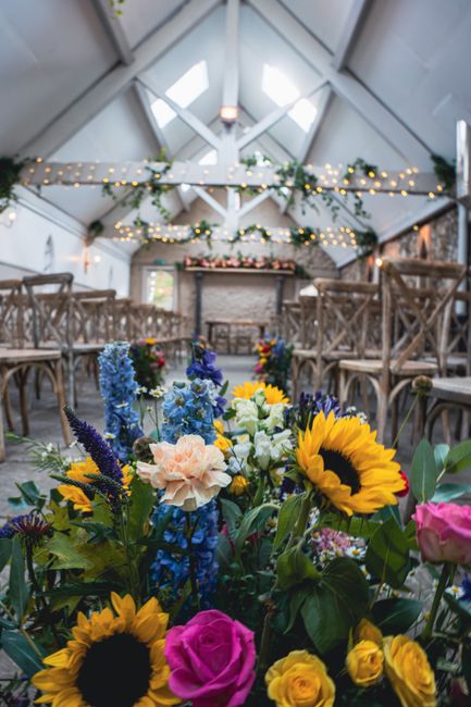 A vibrant floral arrangement featuring sunflowers, pink roses, delphiniums, and yellow roses in the foreground, arranged in a lush bouquet with green foliage. The bouquet is placed inside a modern, ru