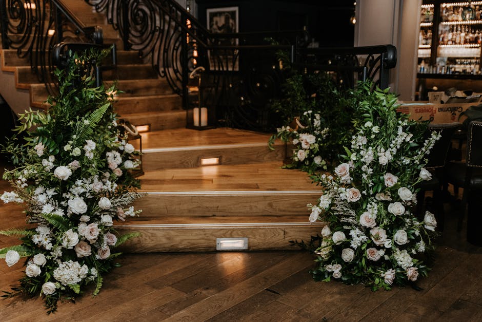 A pair of large floral arrangements featuring white roses, soft pink peonies, and an assortment of greenery including ferns and eucalyptus, positioned on either side of a wooden staircase with built-i