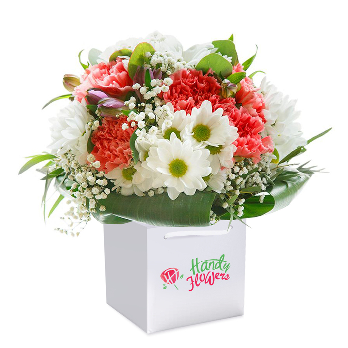 Resplendent in a modern white gift bag, this lush, rounded bouquet is a delightful composition created by Becontree Heath Florist for delivery throughout the local area. Full-bodied carnations in soft coral and fresh peach are loosely interwoven with deeper, passionate reds, their ruffled petals creating a gentle wave of visual texture. These central blooms are animated by the crisp presence of white daisies, each with a lime-green centre that sparkles against the richer colours, reminiscent of wildflowers blooming in the gardens of Becontree Heath. Airy baby's breath, its clusters of petite white blossoms floating gracefully, fills the spaces between with a light and delicate touch. Sprouting softly from within the bouquet, a few closed buds tinged in purple and pink hues promise the magic of blossoms yet to appear. The flowers are encircled by robust, glossy deep green leaves, their lushness recalling the foliage seen along Becontree Avenue. The square white gift bag, highlighted by its practical white ribbon handles, presents the arrangement as a ready-to-gift delight. Captured against a luminous white background, every texture, from the softest carnation to the coolest leaf, stands out in vivid detail. Perfect for conveying happiness or celebrating a special moment, this arrangement exemplifies the quality and care of local floral design available in Becontree Heath.