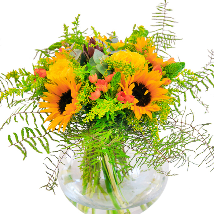The first impression of this bouquet is a sense of pure light and jubilant celebration-a tribute to the sun-drenched avenues of Becontree Heath. Golden yellow roses and striking sunflowers bloom together, their petals unfurling in abundant, generous layers that echo the optimism of summer days in Parsloes Park. Deep green leaves and wispy, airy vines intermingle, cascading from the centre like a verdant waterfall, reminiscent of the lush, shaded paths near the Heathway. Glossy red Hypericum berries sparkle like hidden gems throughout the arrangement, their bold colour boldly punctuating the softer, sunset-hued blooms. Each detail is cradled within a round, luminous glass vase, which accentuates the bouquet's architectural lines and the fresh water quietly nourishing every stem. Up close, there is a dance of textures-from the plushness of roses to the crisp snap of fern sprouts-while the bouquet's gentle fragrance whispers a subtle mix of garden freshness and woodland calm. Perfect for marking joyful milestones, congratulating loved ones, or simply bringing a touch of brightness to the heart of Becontree Heath, this arrangement is a heartfelt gesture from Becontree Heath Florist, reflecting the beauty of our local surroundings.